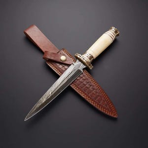 May include: A handcrafted dagger with a patterned steel blade and a bone handle. The dagger rests on a brown leather sheath with a decorative embossed pattern. The sheath has a leather belt loop and a button closure. The dagger's handle has gold-colored accents.