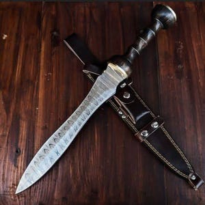 Puede incluir: Vista aérea de una daga con una hoja estampada y un mango de madera oscura, en una funda de cuero. La daga tiene una hoja plateada con un diseño único, y el mango es de color marrón oscuro. La funda es negra con remaches plateados.