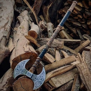May include: A double-bladed axe with a silver blade and intricate Celtic knotwork design. The handle is made of wood and wrapped with dark blue leather. The axe is set against a backdrop of stacked firewood.
