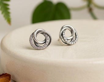 May include: A pair of silver stud earrings featuring an interlocking circular design. Each earring consists of two intertwined rings, with one ring polished smooth and the other ring encrusted with small, clear, sparkling stones along the outer edge.