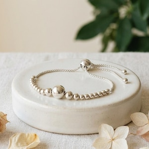 May include: A silver-toned bracelet featuring a series of small metallic beads and one larger central bead. The bracelet includes an adjustable sliding mechanism with two thin chains ending in small spherical stoppers for a secure fit.