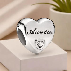 May include: A polished silver heart charm with the word "Auntie" inscribed in cursive. A small heart-shaped gem is set below the word. The charm is designed to be attached to a bracelet or necklace.