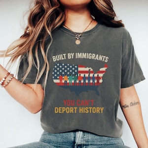 May include: Dark grey t-shirt featuring the text "BUILT BY IMMIGRANTS" above a graphic of the United States map filled with the American flag and silhouettes of people. The text "YOU CAN'T DEPORT HISTORY" is below the map.