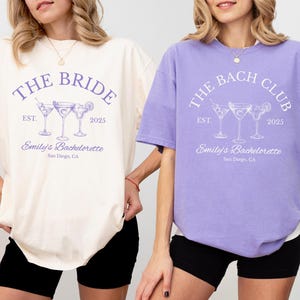 May include: Two short-sleeved t-shirts, one ivory and one violet, with matching text and cocktail graphics. The ivory shirt reads "THE BRIDE" and the violet shirt reads "THE BACH CLUB". Both shirts say "Emily's Bachelorette, San Diego, CA".