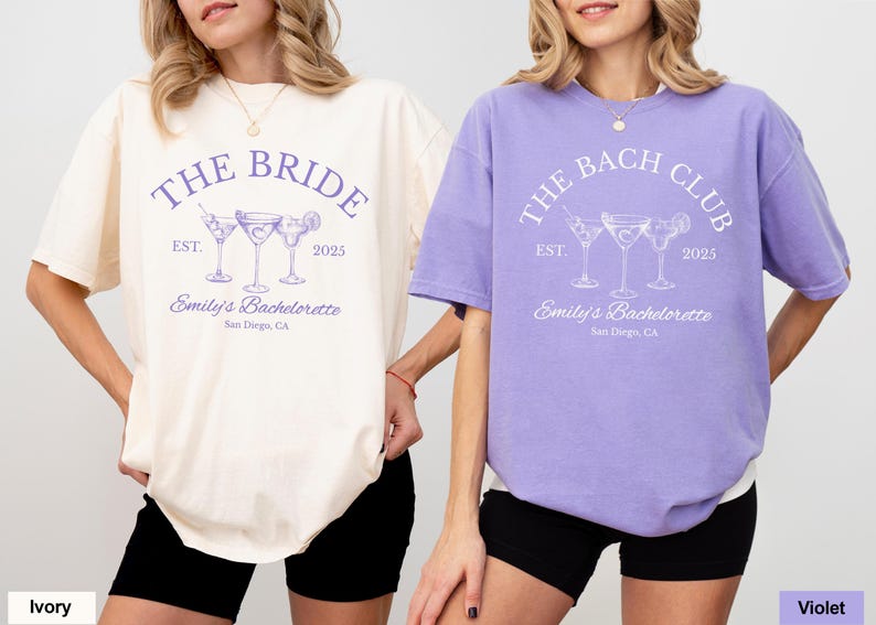 May include: Two short-sleeved t-shirts, one ivory and one violet, with matching text and cocktail graphics. The ivory shirt reads "THE BRIDE" and the violet shirt reads "THE BACH CLUB". Both shirts say "Emily's Bachelorette, San Diego, CA".