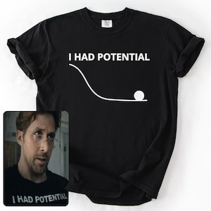 May include: Black t-shirt with the phrase "I HAD POTENTIAL" in white, along with a graphic of a ball rolling down a curved path. The shirt also features a small image of a person wearing a similar shirt.
