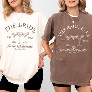 May include: Two t-shirts, one ivory and one espresso, featuring a cocktail design. The ivory shirt reads "THE BRIDE" with "Emily's Bachelorette" and "San Diego, CA." The espresso shirt says "THE BACH CLUB."