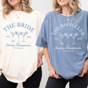 May include: Two short-sleeved t-shirts, one ivory and one blue, with matching text and graphics. The ivory shirt reads "THE BRIDE" above three cocktail glasses, and the blue shirt reads "THE BACH CLUB". Both shirts include "Emily's Bachelorette, San Diego, CA" and the year 2025.