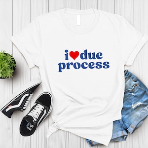 May include: White t-shirt with the text "I ❤️ due process" in blue and a red heart. The shirt is styled with denim shorts and black and white sneakers. The shirt is laying on a white wooden surface.
