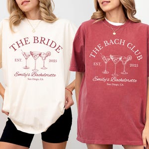 May include: Ivory and crimson-colored t-shirts with the text "THE BRIDE" and "THE BACH CLUB" printed on them. Each shirt features cocktail glasses, the year "2025", and "Emily's Bachelorette, San Diego, CA".