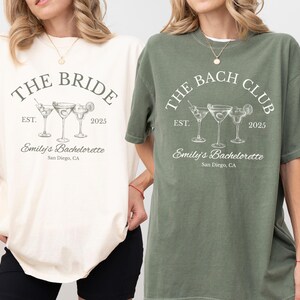 May include: Two short-sleeved t-shirts, one ivory and one moss green, featuring a cocktail-themed design. The ivory shirt reads "THE BRIDE" with "Emily's Bachelorette, San Diego, CA" below. The green shirt says "THE BACH CLUB" with the same details.