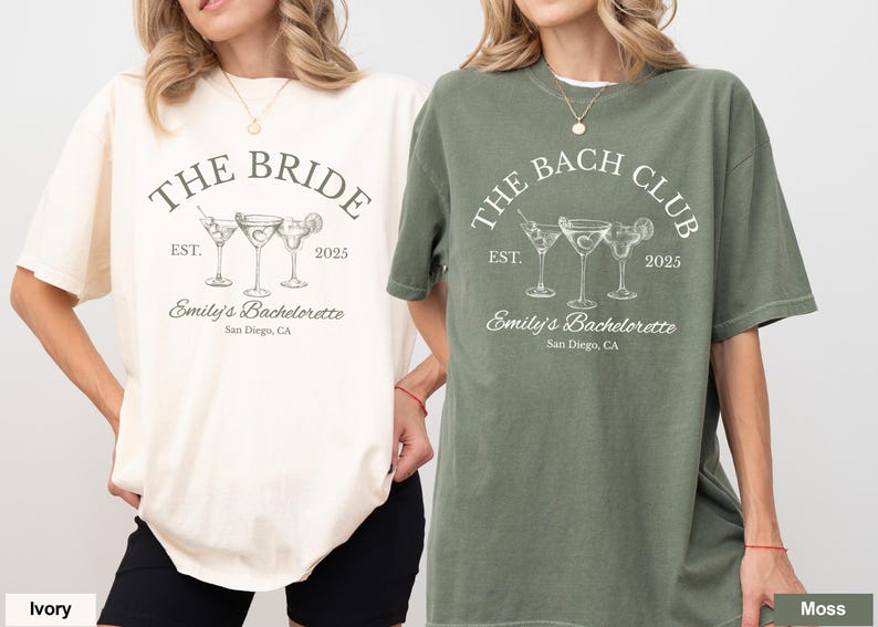 May include: Two short-sleeved t-shirts, one ivory and one moss green, featuring a cocktail-themed design. The ivory shirt reads "THE BRIDE" with "Emily's Bachelorette, San Diego, CA" below. The green shirt says "THE BACH CLUB" with the same details.
