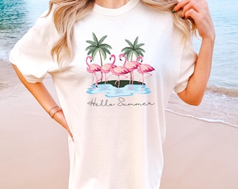 May include: A cream-coloured t-shirt with a graphic of pink flamingos, palm trees, and the text "Hello Summer." The design is set against a blue water background. The shirt has short sleeves and a relaxed fit.