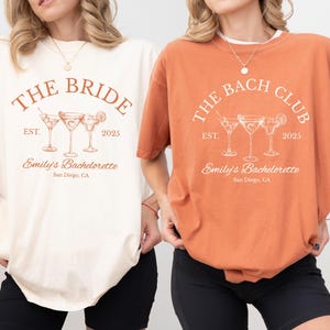 May include: Two t-shirts, one ivory and one yam-colored, with a cocktail-themed design. The ivory shirt reads "THE BRIDE" above three cocktail glasses, "EST. 2025" and "Emily's Bachelorette, San Diego, CA." The yam shirt says "THE BACH CLUB" with the same design.