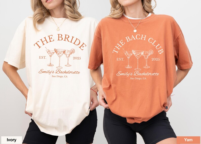 May include: Two t-shirts, one ivory and one yam-colored, with a cocktail-themed design. The ivory shirt reads "THE BRIDE" above three cocktail glasses, "EST. 2025" and "Emily's Bachelorette, San Diego, CA." The yam shirt says "THE BACH CLUB" with the same design.
