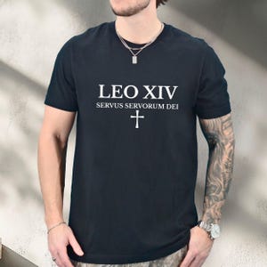 May include: Black t-shirt with white text that reads "LEO XIV SERVUS SERVORUM DEI" above a white cross. The shirt is short-sleeved and has a crew neck. The shirt is worn by a person.