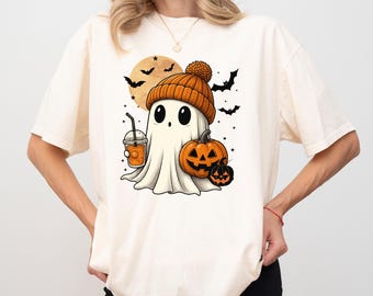 May include: Cream-colored t-shirt featuring a Halloween-themed graphic. The design includes a cartoon ghost wearing an orange beanie, holding a drink, and accompanied by pumpkins and bats. A full moon is in the background.
