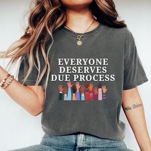 May include: Dark gray t-shirt with the text "EVERYONE DESERVES DUE PROCESS" in white. Below the text are illustrations of hands with various skin tones and clothing patterns. The shirt is a casual, unisex style.