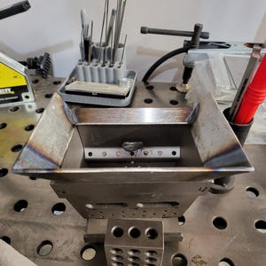 以下が含まれることがあります： 角度の付いた側面を持つ正方形の容器を備えた、製造された金属溶接プロジェクト。 容器は、溶接マグネットや赤いマーカーなど、さまざまなツールや機器を備えた金属表面に置かれています。