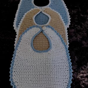Puede incluir: Tres baberos de bebé hechos a mano con ganchillo en blanco, beige y azul claro. Cada babero tiene un borde festoneado y un cierre de botón en el cuello. Los baberos están apilados, mostrando los diferentes colores y texturas del ganchillo.