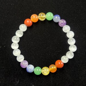 May include: A beaded bracelet featuring a variety of colorful gemstones and white beads. The gemstones are arranged in a rainbow pattern, including red, orange, yellow, green, blue, and purple. The bracelet is on a black surface.