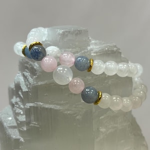 May include: Two beaded bracelets. One bracelet features white, pink, and blue beads with gold accents. The other bracelet is made of white beads. The bracelets are displayed on a white, textured surface.