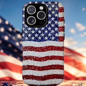 May include: A smartphone case featuring a glittery American flag design with a blue field of white stars in the upper left corner and alternating red and white horizontal stripes. The case has a black camera module cutout with three lenses.