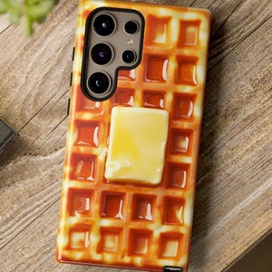May include: A phone case featuring a waffle design with a square of butter on top. The case has a glossy finish and a realistic appearance, with a camera cutout at the top. The case is on a wooden surface.