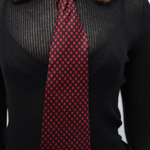 May include: A black tie with a red polka dot pattern is knotted at the neck. The tie is worn over a black long-sleeved top. The image is a close-up of the tie and top.