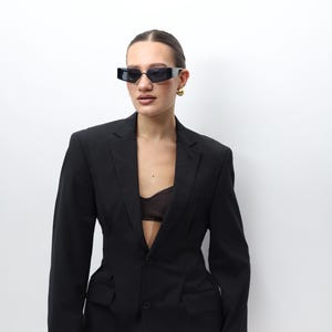 May include: A black blazer with a pinstripe design, featuring a tailored fit and two front pockets. The blazer is paired with black rectangular sunglasses and gold earrings. The outfit is set against a white background.