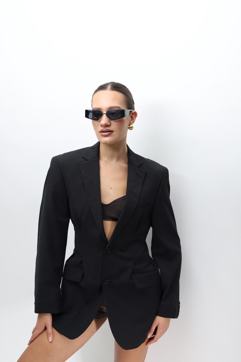 May include: A black blazer with a pinstripe design, featuring a tailored fit and two front pockets. The blazer is paired with black rectangular sunglasses and gold earrings. The outfit is set against a white background.
