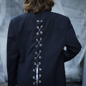Puede incluir: Un blazer negro con un diseño único. La parte trasera del blazer presenta una serie de ojales plateados entrelazados con un cordón gris, creando un patrón de X. Un blazer de estilo clásico con un toque moderno.