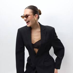 May include: A black blazer with a tailored fit. The blazer features a notched lapel, a single-button closure, and a subtle pinstripe pattern. The model is wearing brown sunglasses and gold earrings.