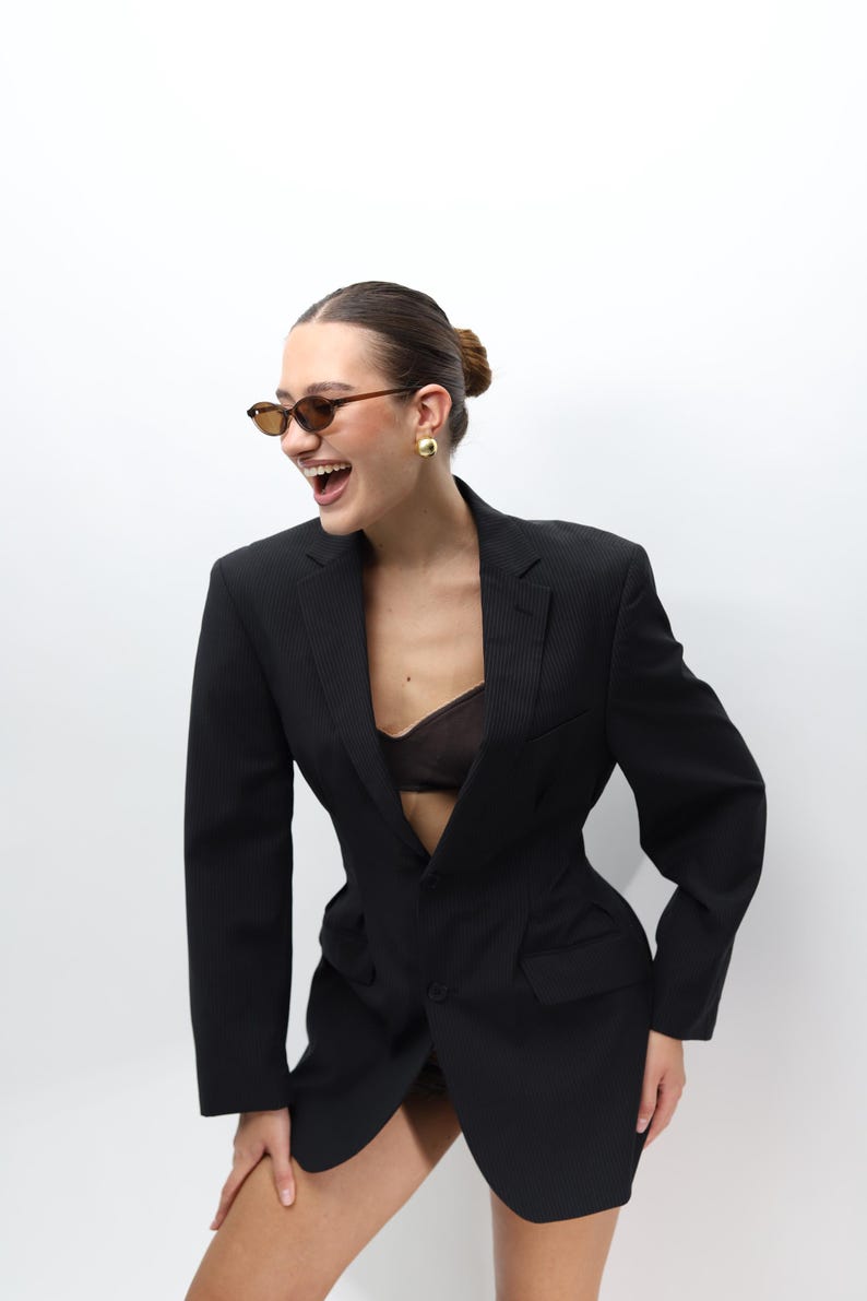 May include: A black blazer with a tailored fit. The blazer features a notched lapel, a single-button closure, and a subtle pinstripe pattern. The model is wearing brown sunglasses and gold earrings.