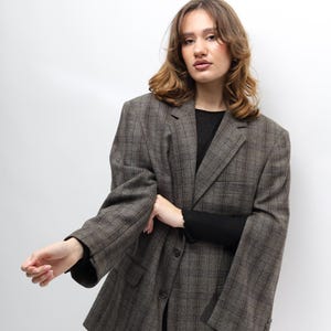 May include: A brown plaid blazer with a classic design. The blazer has a notched lapel, two front pockets, and a two-button closure. The sleeves are long and the fit is oversized. The model is wearing a black top and black trousers.