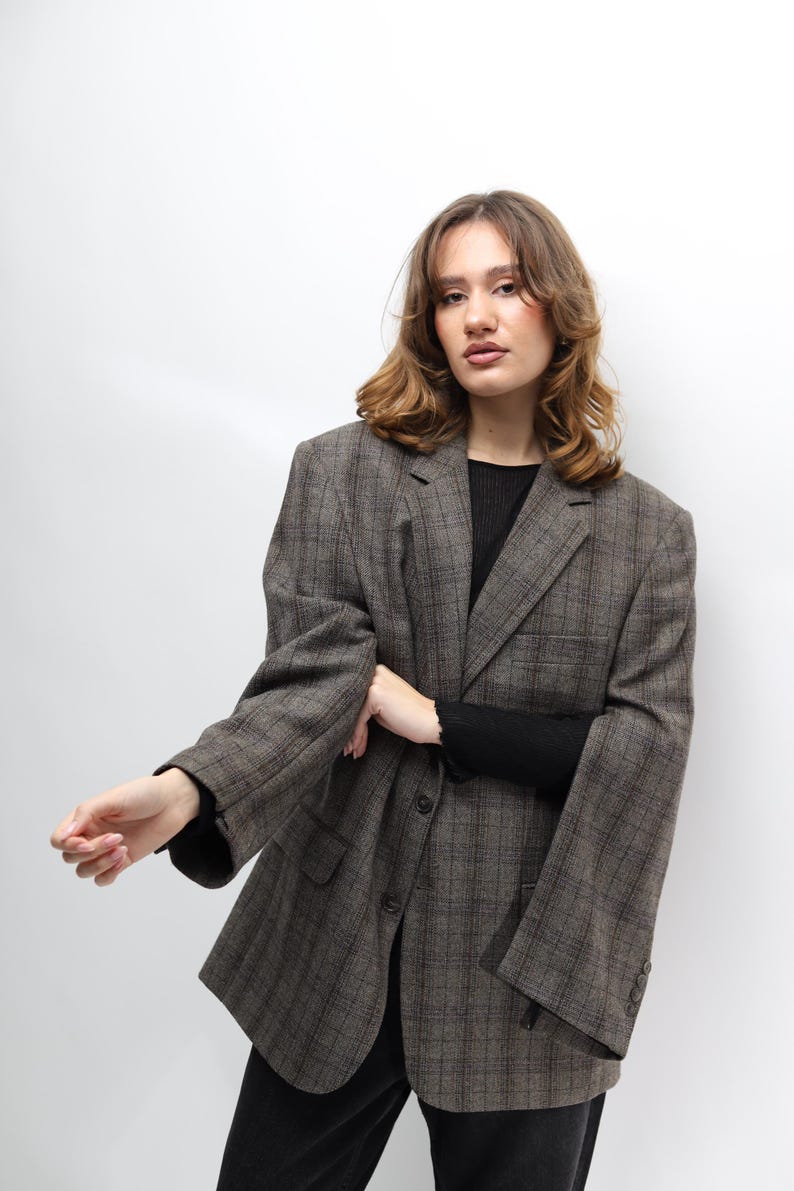 May include: A brown plaid blazer with a classic design. The blazer has a notched lapel, two front pockets, and a two-button closure. The sleeves are long and the fit is oversized. The model is wearing a black top and black trousers.