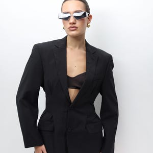 May include: A black pinstripe blazer with a tailored fit. The blazer features a single-button closure, two front pockets, and a notched lapel. The model is wearing futuristic silver sunglasses and gold earrings.