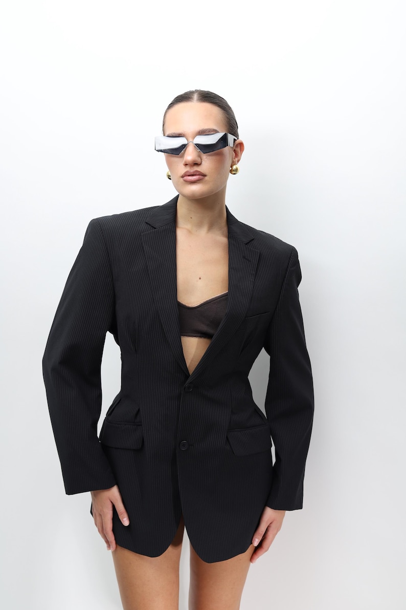 May include: A black pinstripe blazer with a tailored fit. The blazer features a single-button closure, two front pockets, and a notched lapel. The model is wearing futuristic silver sunglasses and gold earrings.