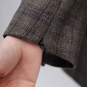 May include: Close-up of a tweed jacket sleeve in shades of brown, grey, and black. The fabric has a textured appearance. A hand is visible near the sleeve's cuff.