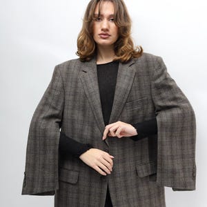 May include: A brown and grey checked blazer with a classic design. The blazer has a notched lapel, two front pockets, and a button closure. The sleeves are long and wide. The person is wearing a black top and black trousers.