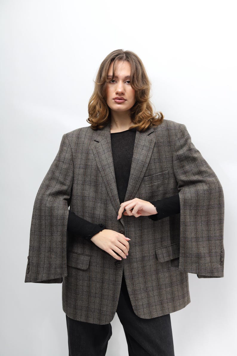 May include: A brown and grey checked blazer with a classic design. The blazer has a notched lapel, two front pockets, and a button closure. The sleeves are long and wide. The person is wearing a black top and black trousers.