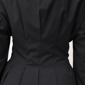 May include: Close-up of a black pinstripe blazer. The blazer has a tailored fit with vertical white stripes. The back of the blazer is visible, showcasing the design details, including a fitted waist and pleats.