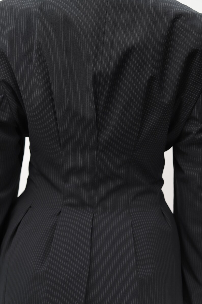 May include: Close-up of a black pinstripe blazer. The blazer has a tailored fit with vertical white stripes. The back of the blazer is visible, showcasing the design details, including a fitted waist and pleats.