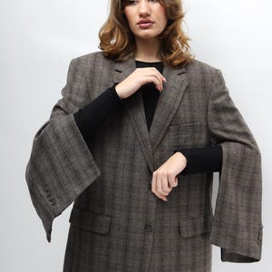 May include: A brown checked blazer with a single-button fastening and two front pockets. The blazer features wide sleeves and a classic notched lapel. The fabric appears to be a wool blend. The model is wearing a black long-sleeved top.