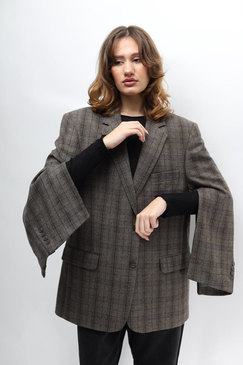 May include: A brown checked blazer with a single-button fastening and two front pockets. The blazer features wide sleeves and a classic notched lapel. The fabric appears to be a wool blend. The model is wearing a black long-sleeved top.