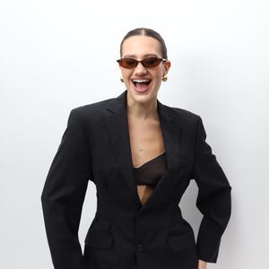May include: A black blazer with a single-button closure and a tailored fit. The blazer has subtle vertical stripes and is worn with a sheer top. The person is wearing brown sunglasses and gold earrings.
