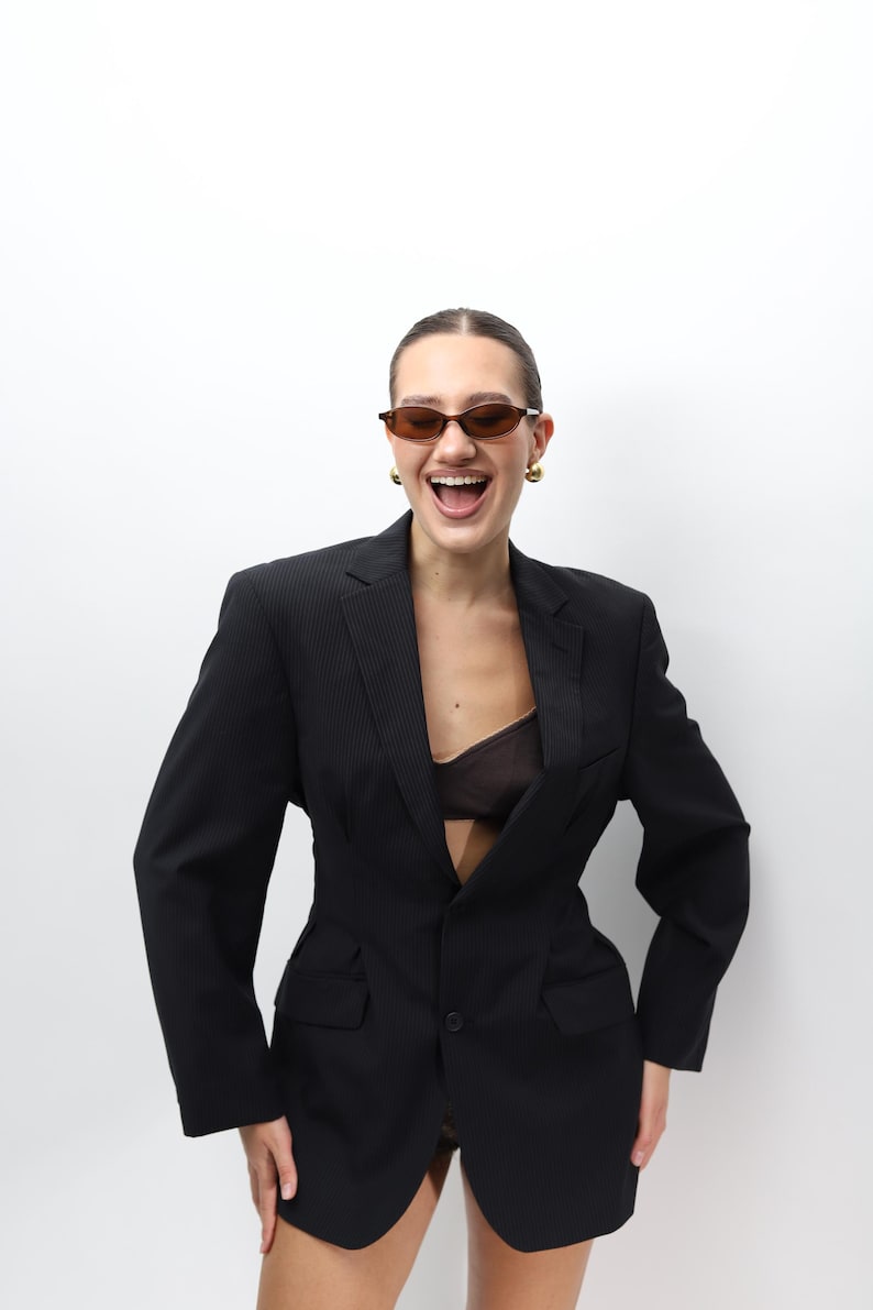 May include: A black blazer with a single-button closure and a tailored fit. The blazer has subtle vertical stripes and is worn with a sheer top. The person is wearing brown sunglasses and gold earrings.
