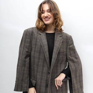 May include: A brown and grey checked blazer with a classic design. The blazer features a notched lapel, a two-button fastening, and a single chest pocket. The sleeves are long and the fabric appears to be woven. The blazer is worn over a black top.