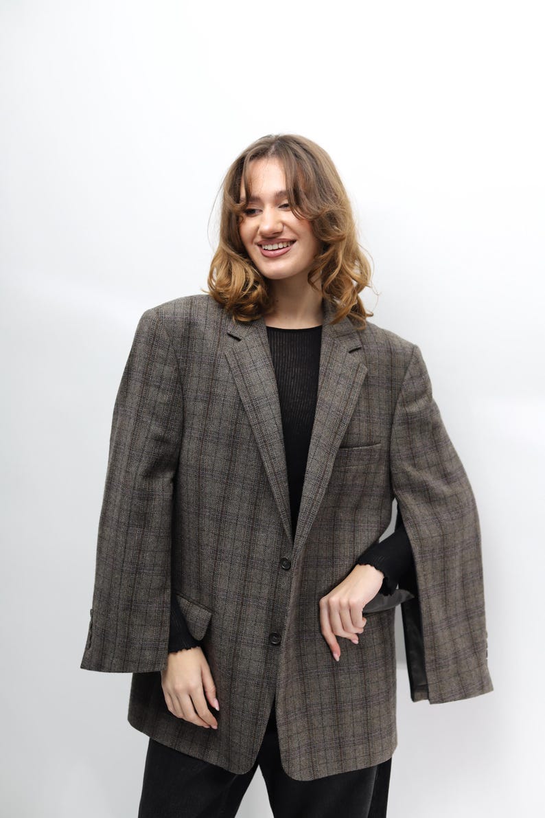 May include: A brown and grey checked blazer with a classic design. The blazer features a notched lapel, a two-button fastening, and a single chest pocket. The sleeves are long and the fabric appears to be woven. The blazer is worn over a black top.