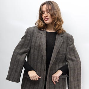 May include: A brown plaid blazer with a classic design. The blazer features a notched lapel, two front pockets, and button closures. The model is wearing a black long-sleeve top and dark jeans. A versatile piece for any wardrobe.